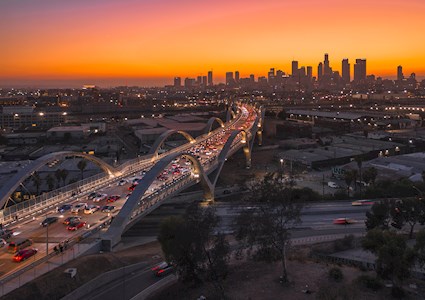 A four-lane bridge with arches stands above a Los Angeles highway. The city of LA is in the background, along with an orange sky. 