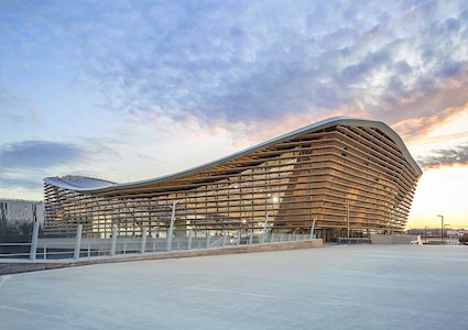 The finished building displays the timber elements comprising the sides and roof. A white pedestrian bridge is seen in the foreground and surrounding buildings in the background. 
