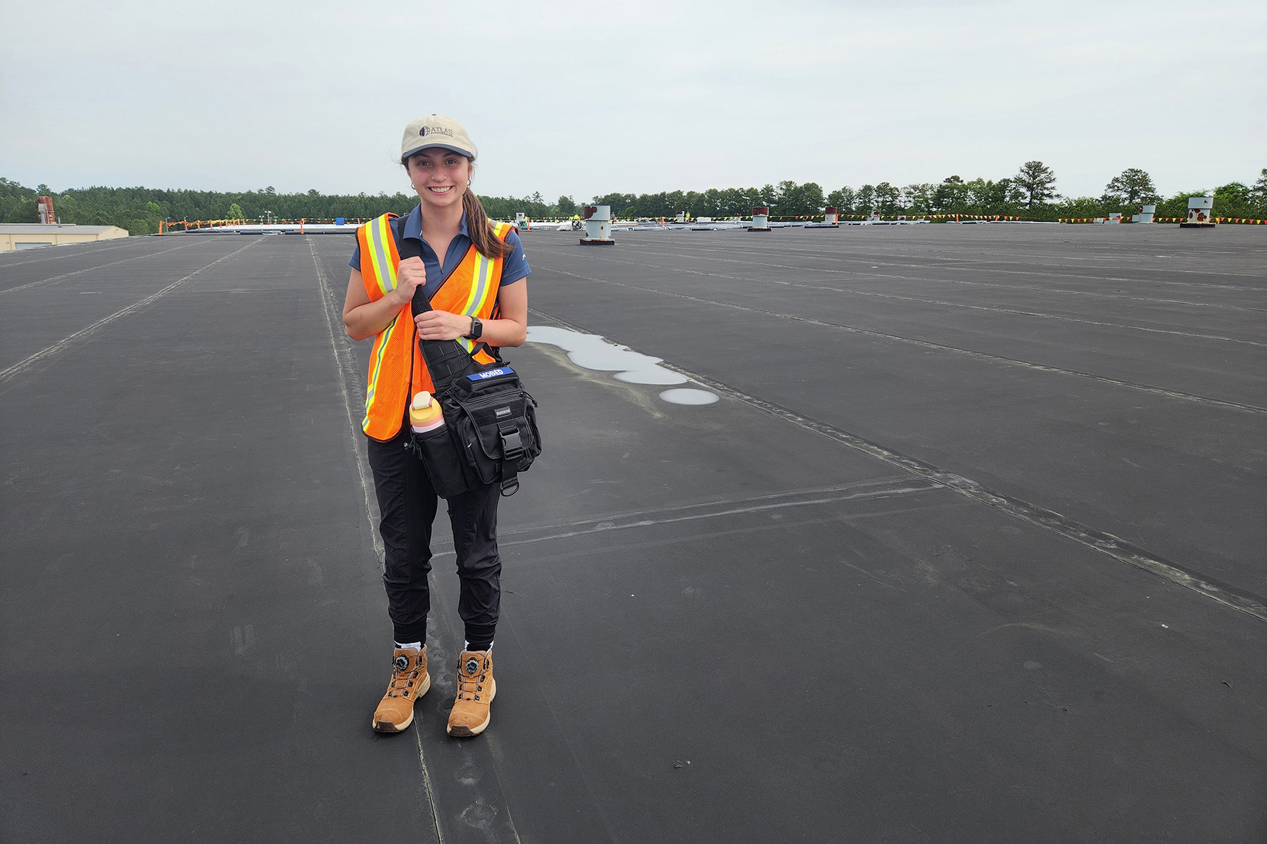 A smiling young woman, in safety vest and Atlas cap, stands atop the roof of a building.