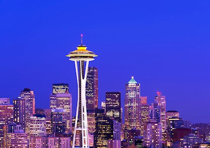 Photograph shows a structure with tall steel columns that taper in near the midsection with a saucer-like top lit up at night and surrounded by other buildings. The columns are connected to a central core. 